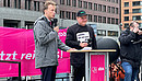 Zwei junge Männer stehen auf einem Podest auf einer Demonstration, der linke der beiden, Florian Schütz, spricht in ein Mikrofon. 