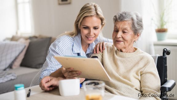 Tag der pflegenden Angehörigen Eine junge Frau sitzt mit einer älteren Frau im Rollstuhl am Esstisch und hilft ihr am Tablet.