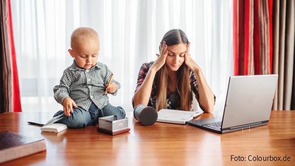 Eine sichtlich gestresste Mutter versucht zu Hause am Laptop zu arbeiten, während neben ihr auf dem Schreibtisch ein Kleinkind sitzt und mit Büroutensilien spielt.