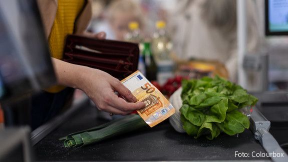An einer Supermarktkasse reicht eine Hand einen Geldschein Richtung Kasse, um zu bezahlen. Auf dem Kassenband liegen Lebensmittel.