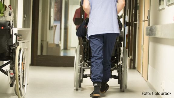 Eine Pflegekraft, von hinten zu sehen, schiebt einen älterne Menschen im Rollstuhl über einen Flur .