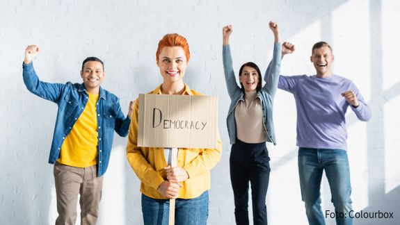 smiling woman holding placard with democracy lettering near multicultural like-minded people showing win gesture on blurred background