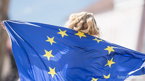 Das Foto zeigt ein Mädchen, das eine EU-Fahne als Umgang nutzt. Die Sterne sind zu erkennen, über dem Rand der Fahne ihre Haare.