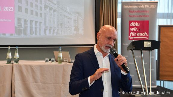 Ein Mann steht vor einer Leinwand mit einem Mikrofon in der Hand 