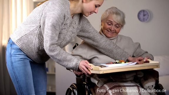 Eine junge Frau reicht einer älteren Frau im Rollstuhl ein Tablett mit einem Mittagessen.