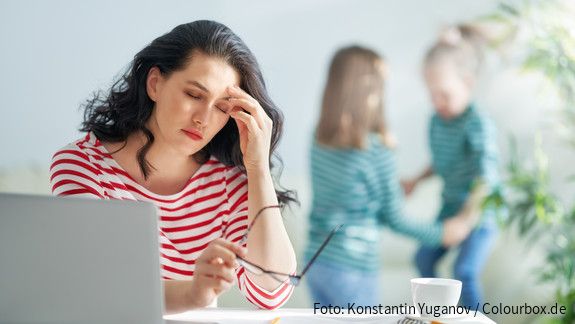 Sorgearbeit fair teilen Eine dunkelhaarige Frau arbeitet am Laptop und reibt sich gestresst die Augen, im Hintergrund spielen zwei kleine Mädchen