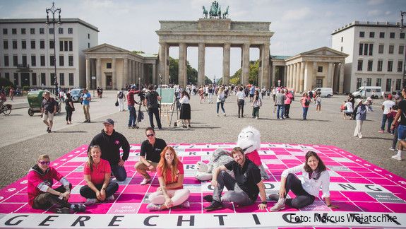 Junge Menschen sitzen vor dem Brandenburger Tor in Berlin auf einem überdimensionierten Scrabble-Spielfeld, auf dem das Wort Gerechtigkeit steht