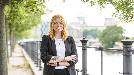 Eine junge Frau steht mit verschränkten Armen draußen und lächelt in die Kamera.