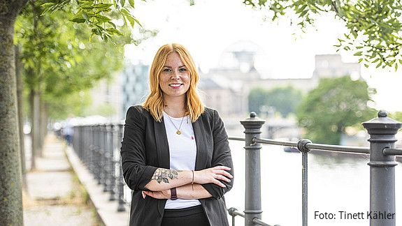 Eine junge Frau steht mit verschränkten Armen draußen und lächelt in die Kamera.