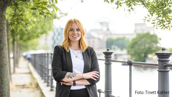 Eine junge Frau steht mit verschränkten Armen draußen und lächelt in die Kamera.