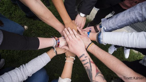 Gesundheitsseminar Nahaufnahme von den Unterarmen junger Menschen, die im Kreis stehen und ihre Hände in der Mitte gemeinschaftlich übereinander legen.