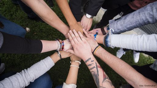 Nahaufnahme von den Unterarmen junger Menschen, die im Kreis stehen und ihre Hände in der Mitte gemeinschaftlich übereinander legen.