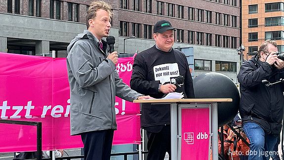 Zwei junge Männer stehen auf einem Podest auf einer Demonstration, der linke der beiden, Florian Schütz, spricht in ein Mikrofon. 