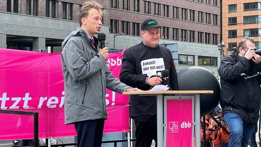 Zwei junge Männer stehen auf einem Podest auf einer Demonstration, der linke der beiden, Florian Schütz, spricht in ein Mikrofon. 