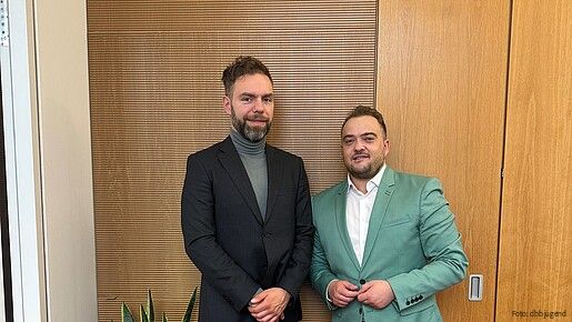 Zwei Männer in Anzügen stehen vor einer Holzwand und blicken in die Kamera