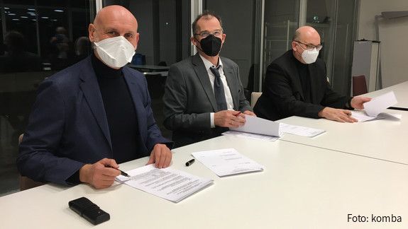 Drei Männer mit Mund-Nasenschutz sitzen an einem Tisch und unterzeichnen die Verhandlungsergebnisse.